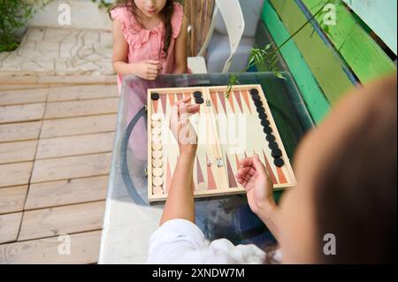 Eine Mutter und ihre Tochter spielen ein lustiges Spiel Backgammon an einem Tisch im Freien. Der Moment der Familienbindung wird in einer natürlichen, sonnigen Umgebung hervorgehoben Stockfoto