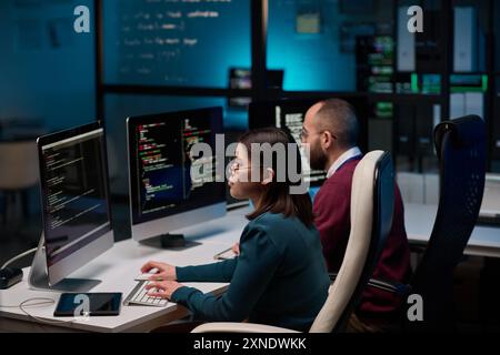 Hochwinkel auf zwei Personen, die am Arbeitsplatz in der Cybersicherheitsabteilung sitzen und Code in blauen Lichtern schreiben Stockfoto