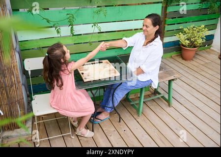 Mutter und Tochter genießen ein lustiges Spiel Backgammon zusammen im Freien. Sie tauschen ein Lächeln und einen Handschlag aus, um Familienbeziehungen und Freizeit hervorzuheben Stockfoto