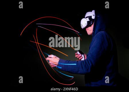 Ein Mann mit Hoodie und Meta Quest 3-Headset taucht in ein gewalttätiges Virtual-Reality-Kampfspiel ein. Lichtmalerei mit langer Belichtung mit Neonlichtern. Stockfoto