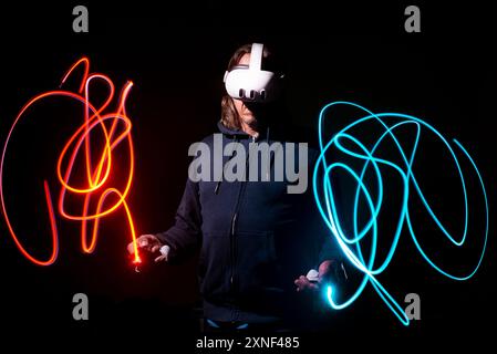 Mann mit einem Meta Quest 3-Headset, eingetaucht in die virtuelle Realität, umgeben von Neonlichtspuren in einem Lichtgemälde mit langer Belichtung. Stockfoto