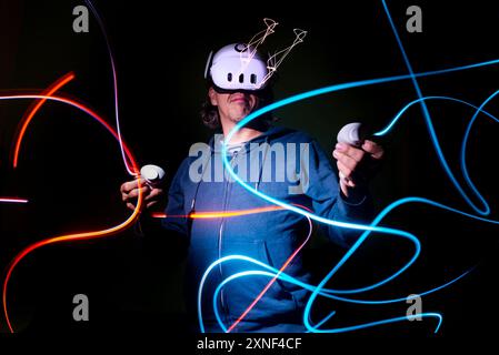 Mann mit einem Meta Quest 3-Headset, eingetaucht in die virtuelle Realität, umgeben von Neonlichtspuren in einem Lichtgemälde mit langer Belichtung. Stockfoto