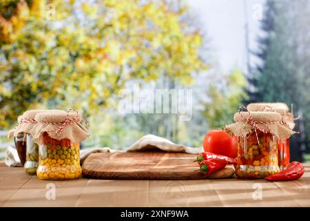 Gläser mit Herbstkonserven von Gemüse und Früchten auf einem Herbsttisch mit einem leeren Platz für Produkte. Im Hintergrund ein Herbstgarten und Bäume Stockfoto