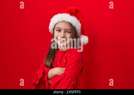 Entzückendes hispanisches Mädchen, Arme gekreuzt, trägt einen weihnachtsmütze über rotem Hintergrund. Ihr fröhliches, strahlendes Lächeln strahlt auf die Kamera. Was für ein Bündel positiver Ergebnisse Stockfoto