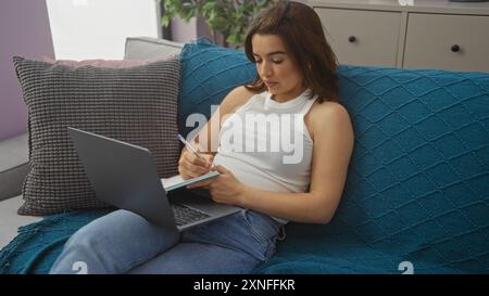 Eine junge hispanische Brünette arbeitet an einem Laptop und macht sich Notizen im Wohnzimmer und präsentiert ein zwangloses Zuhause mit einem komfortablen und modernen ap Stockfoto