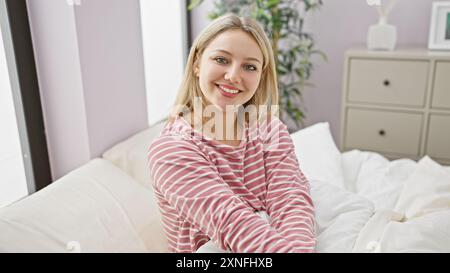 Eine lächelnde blonde Frau im gestreiften Pyjama posiert bequem in einem gemütlichen Schlafzimmerzimmer und strahlt eine entspannte und zugängliche Aura aus. Stockfoto