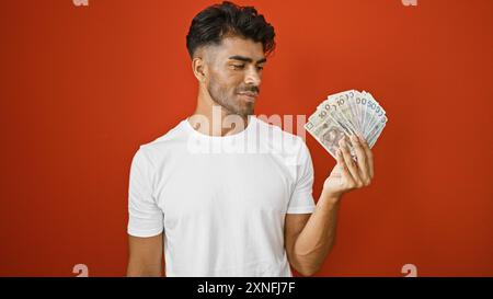 Gutaussehender hispanischer Mann, der polnische Zloty-Banknoten vor rotem Hintergrund untersucht Stockfoto