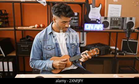 Hispanischer Mann, der in einem Aufnahmestudio Ukulele spielt, mit einem „On Air“-Schild im Hintergrund. Stockfoto