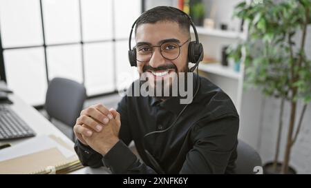 Ein fröhlicher bärtiger Mann, der ein Headset an seinem Schreibtisch in einem hellen, modernen Büro trägt und selbstbewusst lächelt. Stockfoto