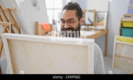 Lächelnder bärtiger Mann mit Brille in einem hellen Kunststudio, der Kreativität und Konzentration ausstrahlt. Stockfoto