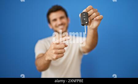 Ein lächelnder Mann, der auf einen Autoschlüssel mit blauem Hintergrund zeigt, der Besitz und Glück darstellt. Stockfoto