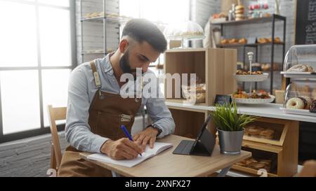 Junger Mann mit Bart, der in einem Notizbuch in einer Bäckerei schreibt, mit verschiedenen Backwaren im Hintergrund Stockfoto