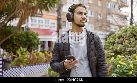 Hübscher hispanischer Mann mit Kopfhörern, der ein Smartphone in einem grünen Stadtpark benutzt. Stockfoto