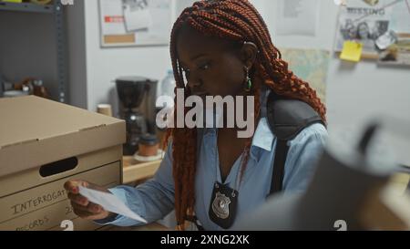 Eine fokussierte afroamerikanische Detektivin untersucht Papierkram in einem überladenen Polizeibüro. Stockfoto