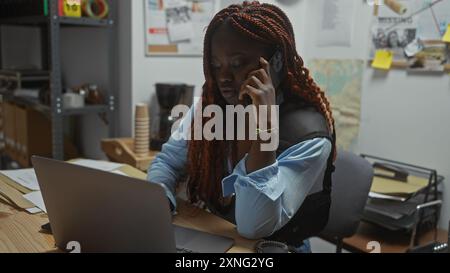 Eine fokussierte afroamerikanische Detektivin, die in einem Polizeirevier arbeitet, einen Laptop benutzt und telefoniert. Stockfoto