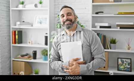 Gutaussehender ranghanischer Mann mit grauem Bart, der im modernen Büro lächelt und Dokumente hält Stockfoto