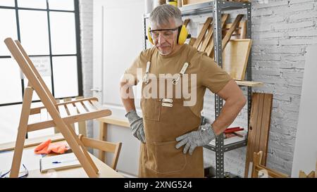 Senior man in der Schürze posiert selbstbewusst in einer gut ausgestatteten Tischlerei und verkörpert Erfahrung und Präzision. Stockfoto