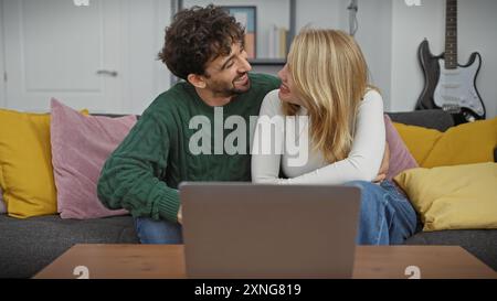 Ein liebevolles Paar teilt sich einen freudigen Moment auf einer bunten Couch in einem gemütlichen Wohnzimmer, während es sich über einen Laptop miteinander verbindet. Stockfoto