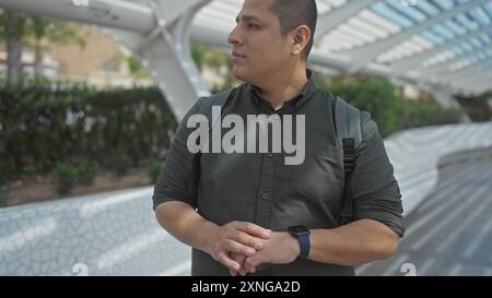 Ein nachdenklicher junger hispanischer Mann mit Smartwatch und Rucksack steht in einem urbanen Park mit moderner Architektur. Stockfoto