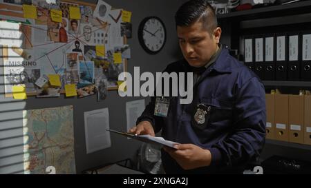 Hispanischer männlicher Detective, der Dokumente im Untersuchungsraum einer Polizeiwache mit Beweisbrett und Uhr analysiert Stockfoto