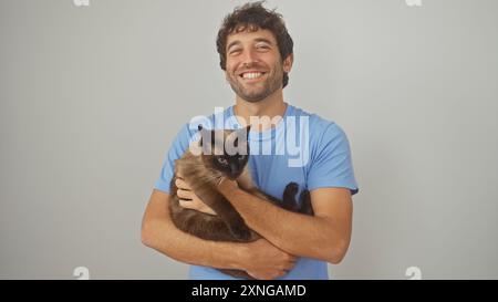 Lächelnder junger erwachsener hispanischer Mann, der eine siamesische Katze vor einem isolierten weißen Hintergrund hält Stockfoto