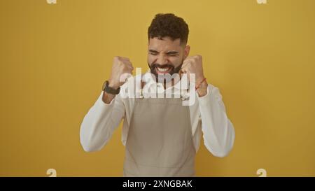 Aufgeregter bärtiger Mann in Schürze, der Fäuste über isoliertem gelbem Hintergrund zusammenballt und Glück und Erfolg zeigt. Stockfoto