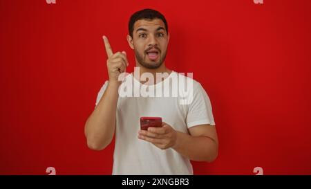 Junger Mann, der das Smartphone hält und den Finger vor rotem Hintergrund hochgehoben hat, einen Punkt betont oder eine Idee mit einem enthusiastischen Ausdruck präsentiert Stockfoto