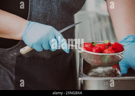 Eine Person, die blaue Handschuhe trägt, wäscht Erdbeeren in einem Metallsieb in einer professionellen Küche. Die Erdbeeren werden in einem rostfreien ste gewaschen Stockfoto