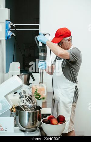Der Konditor trägt eine weiße Schürze und eine rote Kappe und verwendet einen Tauchmixer in einer professionellen Küche, um einen PfirsichSmoothie oder eine Marmelade zuzubereiten Stockfoto