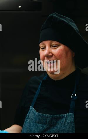 Lächelnde Bäckerin in schwarzem Koch-Outfit und blauer Schürze arbeitet in einer professionellen Küche und zeigt ihre Leidenschaft für die Herstellung köstlicher Backwaren, br Stockfoto