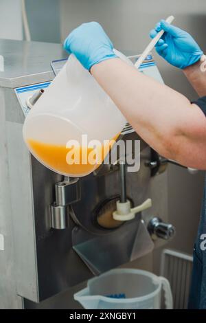 Der Konditor mit blauen Handschuhen gießt eine hausgemachte Sorbet-Mischung aus einem großen Plastikbehälter in einen professionellen Eismacher Stockfoto