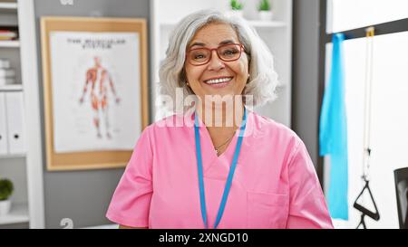 Lächelnde grauhaarige Frau in rosafarbenen Stühlen steht im Krankenhaus mit anatomischem Poster im Hintergrund Stockfoto