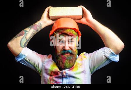 Bärtiger Bauarbeiter im Schutzhelm mit Ziegel. Porträt des lächelnden Maurers im Hardhelm. Mauerwerk. Baumeister Maurer in orange hart Stockfoto