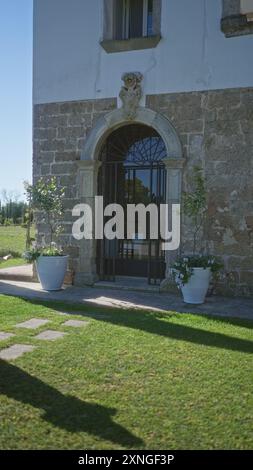 Bezaubernder Eingang aus einem alten Steingebäude in apulien, italien, mit kunstvollen Bogengängen, Topfpflanzen und üppigem grünem Garten unter klarem blauem Himmel. Stockfoto