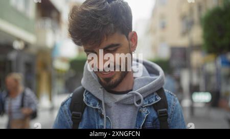 Ein lächelnder junger hispanischer Mann mit Bart, der eine Jeansjacke in der urbanen Stadt trägt. Stockfoto