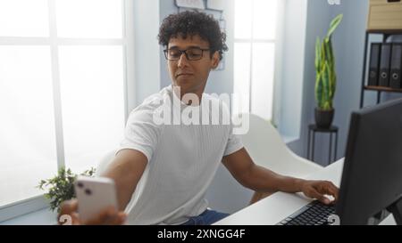 Junger Mann, der im Büro Selfie macht, mit Computer und Pflanze im Hintergrund, mit Brille und weißem Hemd, fokussiert und inhaltlich Stockfoto