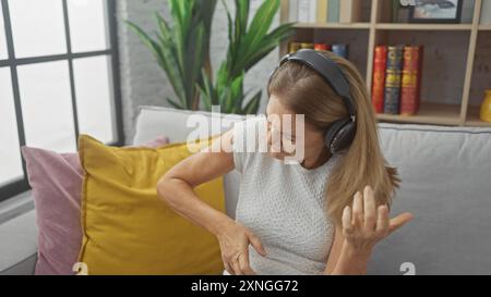 Eine Erwachsene Frau, die Kopfhörer trägt, gibt vor, drinnen Gitarre zu spielen und Musik in einem gemütlichen Wohnzimmer zu genießen. Stockfoto