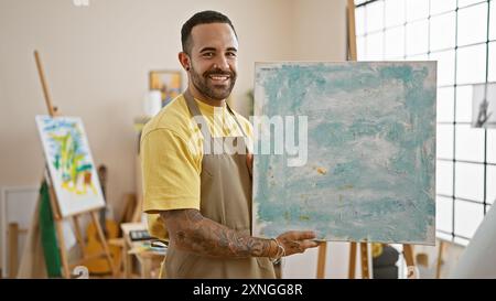 Ein lächelnder bärtiger Mann präsentiert abstrakte Gemälde in einem hellen Atelierinnenraum. Stockfoto