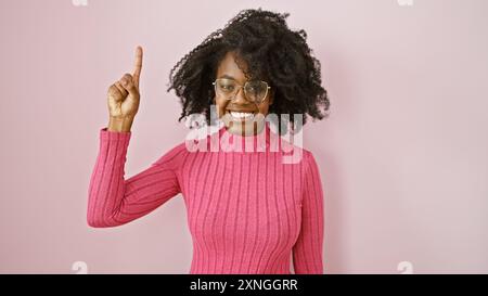 Eine lächelnde afrikanerin mit Brille zeigt nach oben, posiert vor einem rosa Hintergrund in einer ungezwungenen Umgebung. Stockfoto