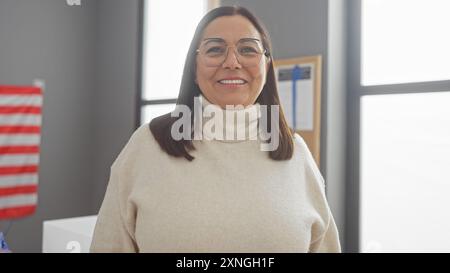 Eine lächelnde hispanische Frau mittleren Alters mit Brille steht drinnen mit einer US-Flagge und Zertifikaten im Hintergrund. Stockfoto