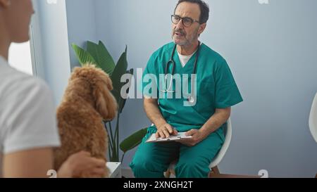 Ein hispanischer Mann in tierarzteppichen berät ein Tablett in einer Klinik und sieht sich einem Pudel und seinem Besitzer gegenüber. Stockfoto