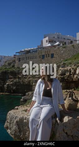 Eine junge, wunderschöne hispanische Frau sitzt auf einem Felsen am Meer in polignano a Mare, apulien, italien, und fängt die ruhige Stranglandschaft unter einem ein Stockfoto