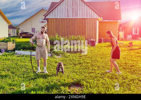 Junge Besitzer eines amerikanischen Bullenhundes haben Spaß mit ihrem Haustier im Hinterhof des Landhauses, Hund trinkt Wasser aus dem Gartenschlauch. Stockfoto