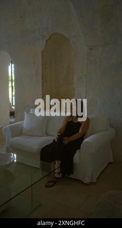 In einem typischen italienischen Haus sitzt eine junge hispanische Frau in einem schwarzen Kleid nachdenklich auf einer weißen Couch vor rustikalen Wänden. Stockfoto