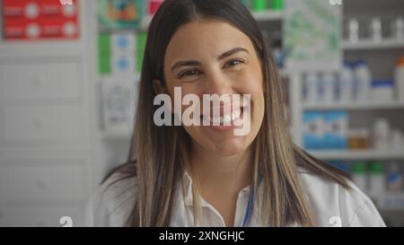 Eine lächelnde junge hispanische Apothekerin steht in einer Drogerie, umgeben von Medikamenten und Gesundheitsvorräten. Stockfoto