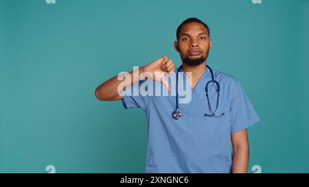 Traurige afroamerikanische Krankenschwester, die während der Arbeit die Daumen nach unten zeigt, isoliert über dem Studio-Hintergrund. Unglücklicher Angestellter im Gesundheitswesen, der Peelings trägt und Mißbilligungsgesten macht, Kamera B Stockfoto