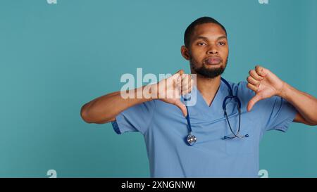 Traurige afroamerikanische Krankenschwester, die während der Arbeit die Daumen nach unten zeigt, isoliert über dem Studio-Hintergrund. Unglücklicher Angestellter im Gesundheitswesen mit Peelings, Mißbilligungsgesten, Kamera A Stockfoto