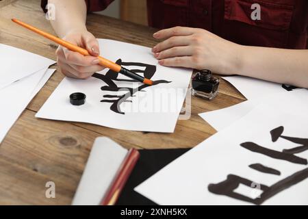 Kalligraphie. Frau mit Pinsel und Tintenfass, die chinesische Hieroglyphen auf Papier auf Holztisch schreibt, Großaufnahme Stockfoto