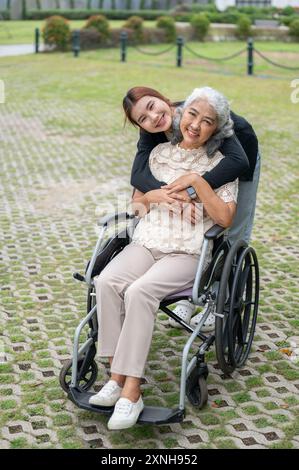Eine hübsche asiatische Enkelin umarmt ihre Oma, die im Rollstuhl sitzt, während sie in einem Park läuft, Zeit miteinander verbringt und sich mit Liebe umschließt Stockfoto