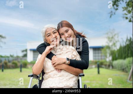 Eine hübsche asiatische Enkelin umarmt ihre Oma, die im Rollstuhl sitzt, während sie in einem Park läuft, Zeit miteinander verbringt und sich mit Liebe umschließt Stockfoto